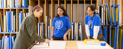 Three people, two wearing RISD tee shirts, are working on a project in a shared RISD workspace.