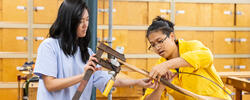 Two students, one in a light purple shirt and one in a yellow jumpsuit, are working together with a large clamp and pieces of wood.