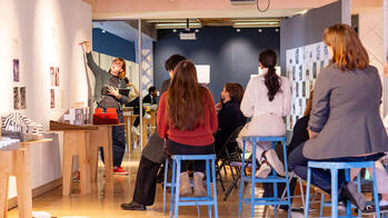 A group of RISD students are gathered on black chairs and blue stools as a fellow student presents their final crit. 