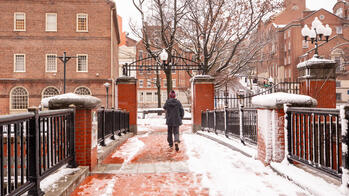 A person wearing a winter coat and boots walks across the College Street bridge in the snow.
