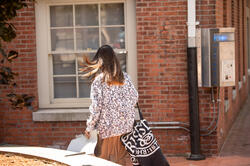 A person with long brown hair wearing a jacket printed with many faces is carrying a RISD tote down a street.