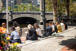 A group of people are sitting along the Providence river outside of RISD’s Canal Walk buildings.